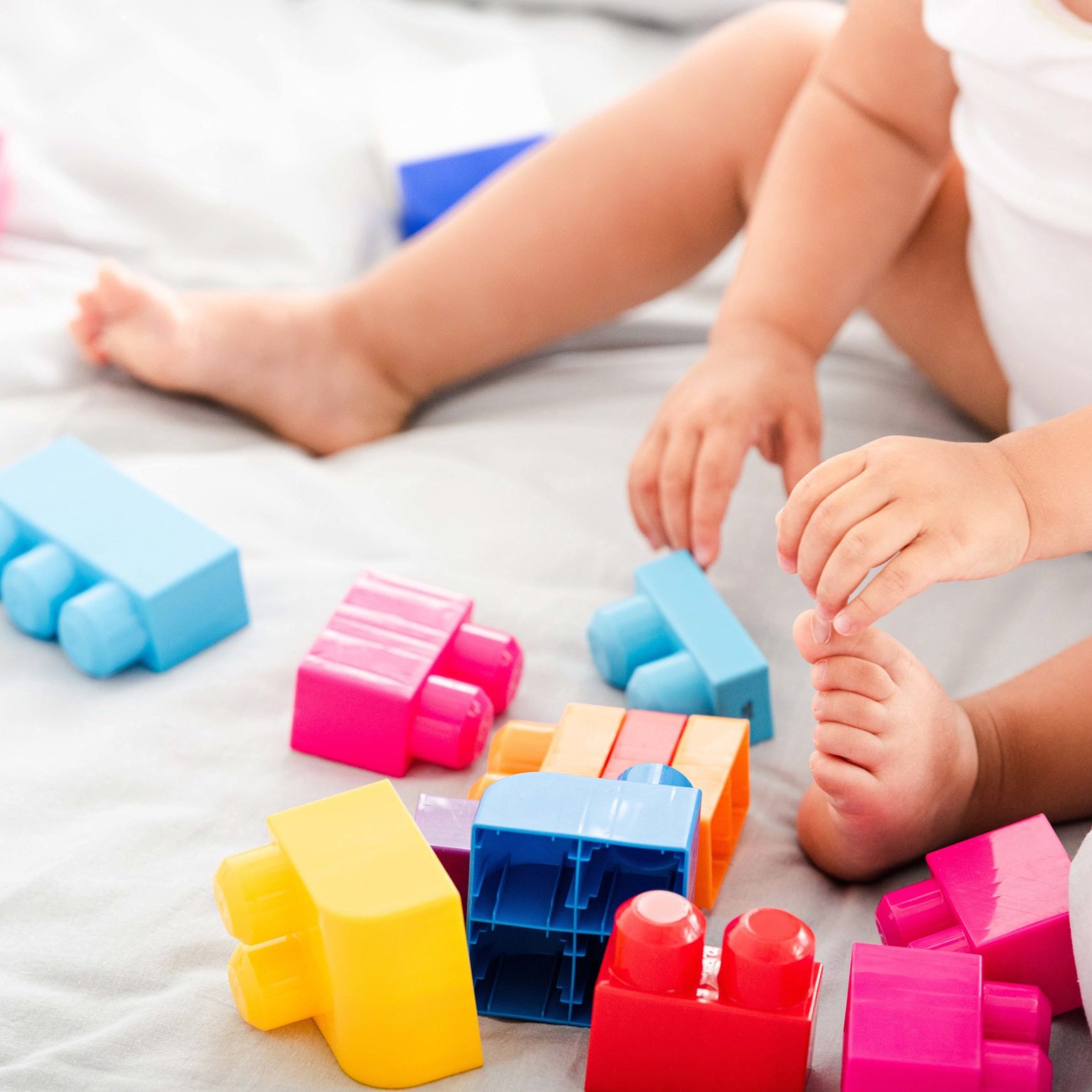 Partial view of barefoot baby in white clothes sitting on bed and playing with bright construction